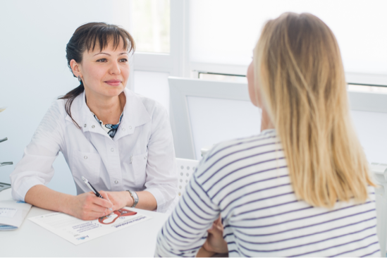 A photograph of a doctor in a white coat writing notes as she seats across from a patient whose back is toward the camera.